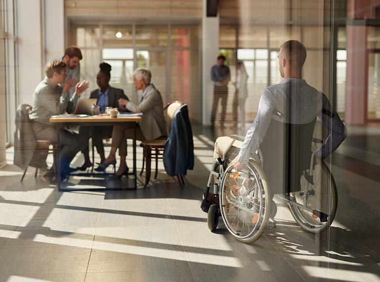 Rear view of a businessman in a wheelchair arriving on a meeting with his colleagues in the office. The view is through glass.