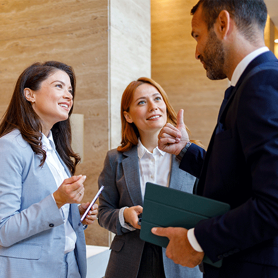Three business professionals in suits having a discussion, with a man gesturing while holding a green folder.