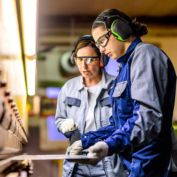 Two workers in safety glasses and ear protection examining materials in an industrial workshop setting.
