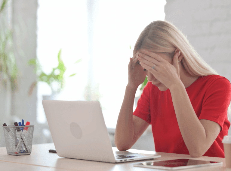Woman in red shirt holding her head in stress while sitting at laptop computer in bright office with plants
