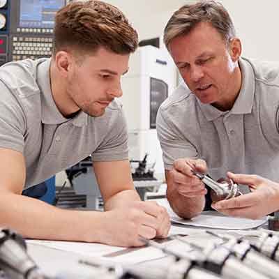 Two men in grey polo shirts reviewing technical documents together in a workshop with machinery in the background.