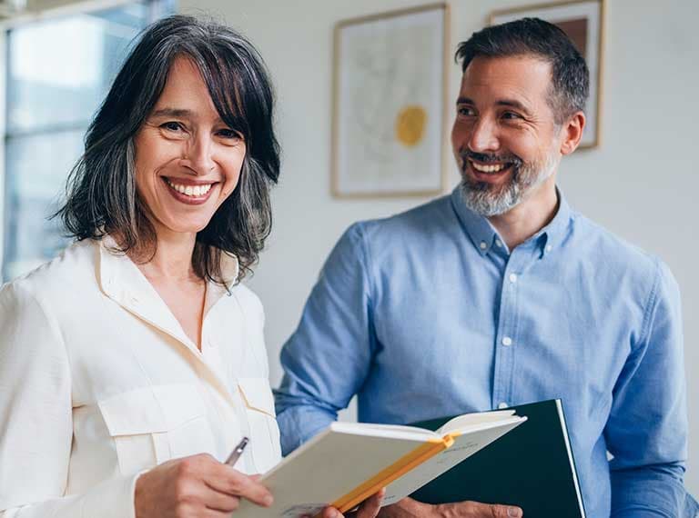 Smiling woman in white shirt holding notebook with pen, standing beside man in blue shirt in bright office setting.
