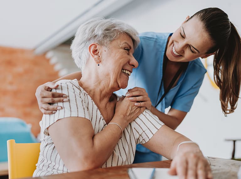Young female caregiver in blue scrubs smiling while assisting a happy senior woman with short gray hair indoors.
