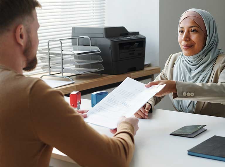 Woman in gray hijab and beige blazer hands document to man across white desk in bright office with printer in background.