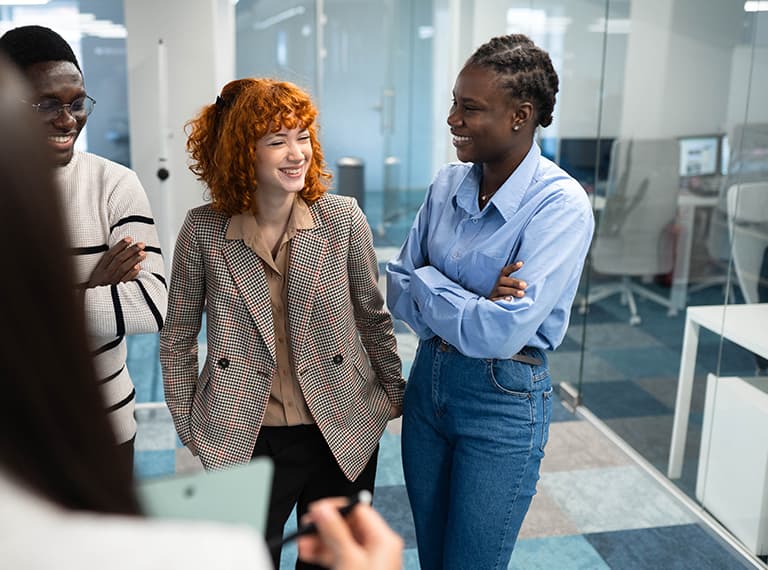 Three diverse professionals smiling and conversing in a modern office with glass walls and bright natural lighting.