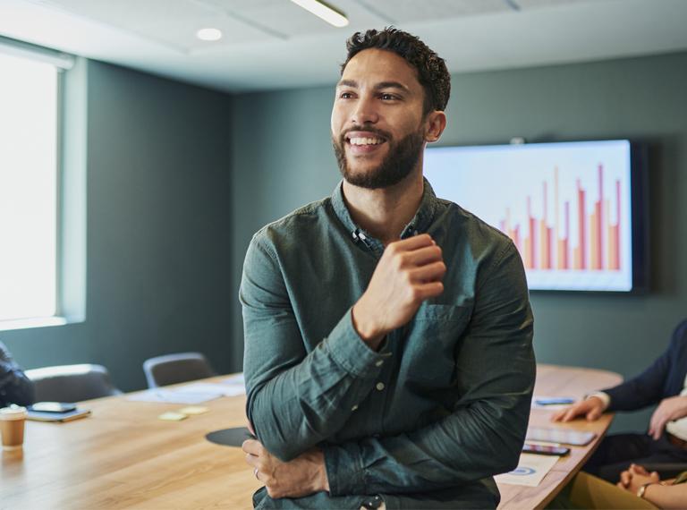 Confident young businessman standing in a modern office and smiling. He is wearing a casual shirt and has a beard. In the background, there are people sitting at a conference table.