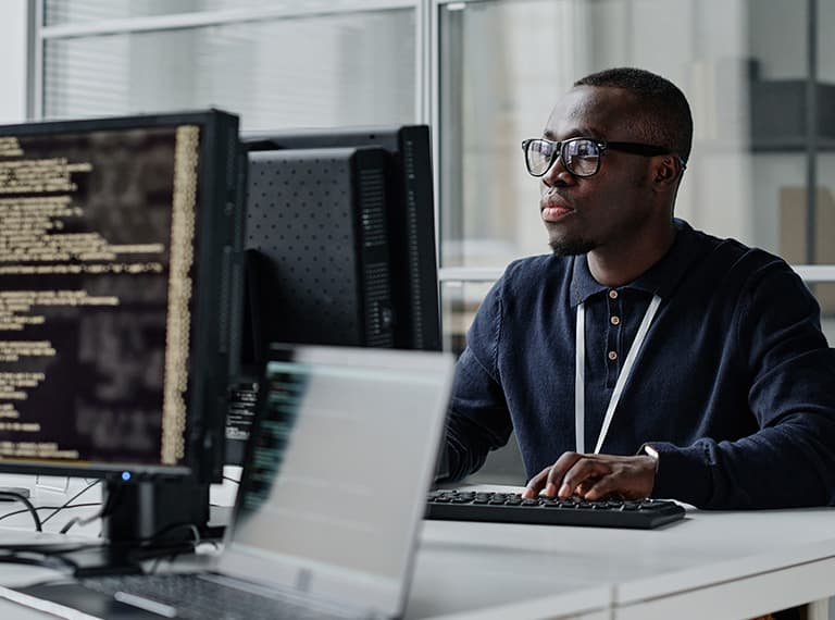 Professional man wearing glasses working at computer with code displayed on multiple monitors in modern office setting