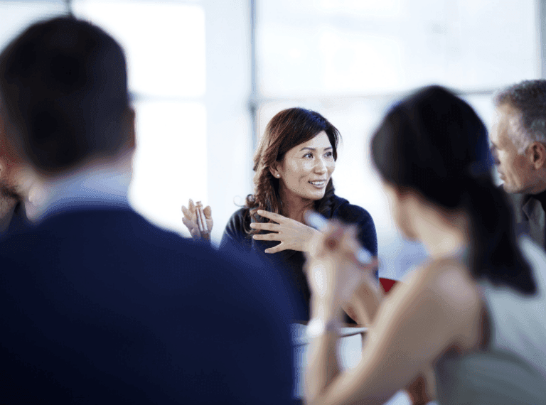Woman with brown hair gesturing while speaking to a group of people in a bright office setting with windows.
