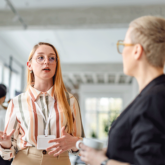 Two professional women having a conversation in a bright office, one wearing glasses and a striped shirt, the other in black.