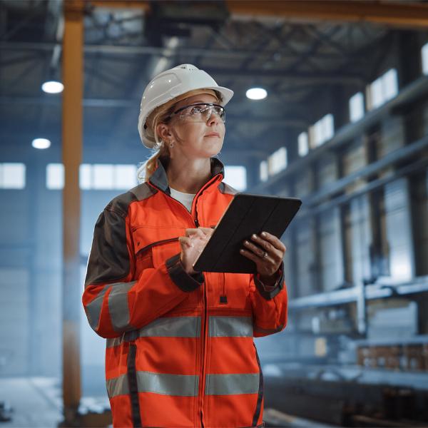 Professional Heavy Industry Engineer Worker Wearing Safety Uniform and Hard Hat, Using Tablet Computer. Serious Successful Female Industrial Specialist Walking in a Metal Manufacture Warehouse.