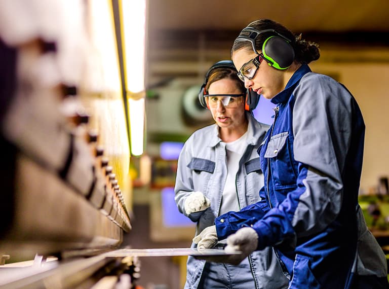 Two workers in safety gear and ear protection examining materials in an industrial workshop setting.
