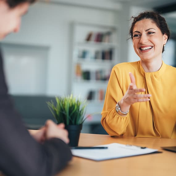 Woman in yellow blazer smiling and gesturing while speaking with colleague in modern office setting with shelves in background.