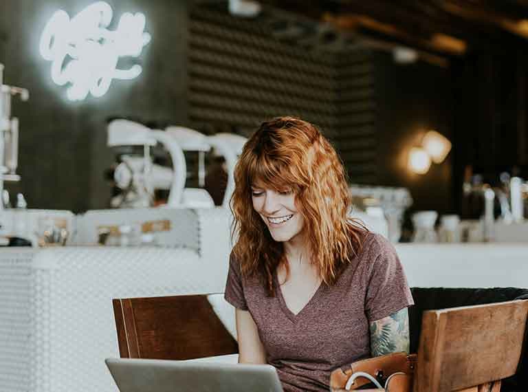 Smiling woman with red hair working on laptop in modern coffee shop with neon sign and wooden chairs.
