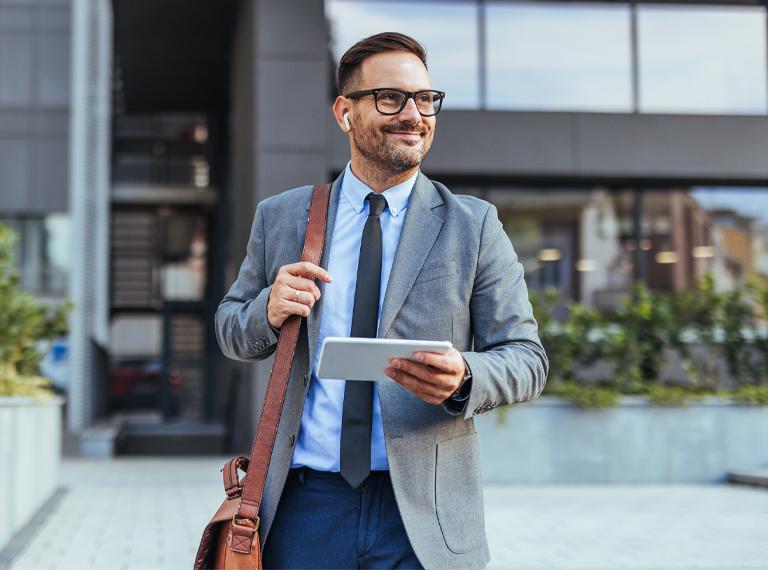Smiling businessman in a suit and tie walking outside a modern office building, holding a tablet. He appears confident and professional, suggesting a successful business environment.