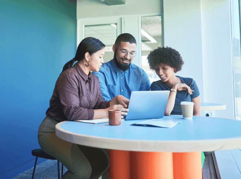 Three diverse colleagues collaborating around a laptop at a blue round table in a modern office with coffee cups.