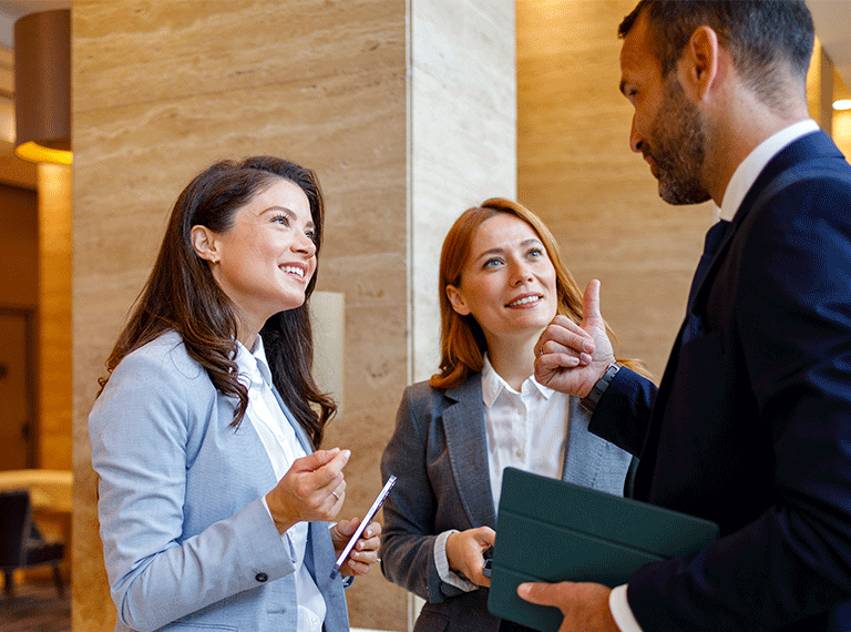 Three business professionals in suits conversing in a modern office lobby with warm lighting and stone walls.