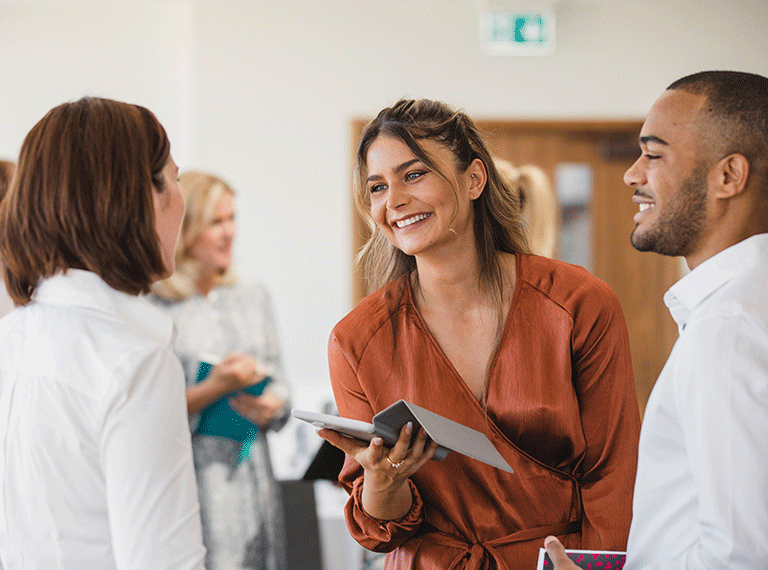 Woman in rust-colored blouse smiling while holding tablet, conversing with colleagues in bright office setting.
