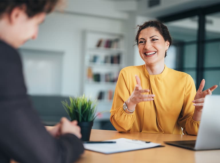 Woman in yellow blouse smiling and gesturing while speaking with colleague in modern office setting.