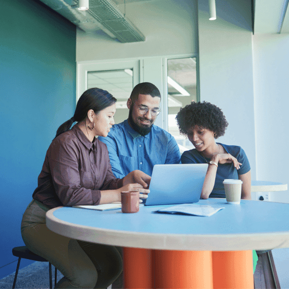 Three diverse colleagues collaborating around a laptop at a round table in a modern office with blue walls and coffee cups.