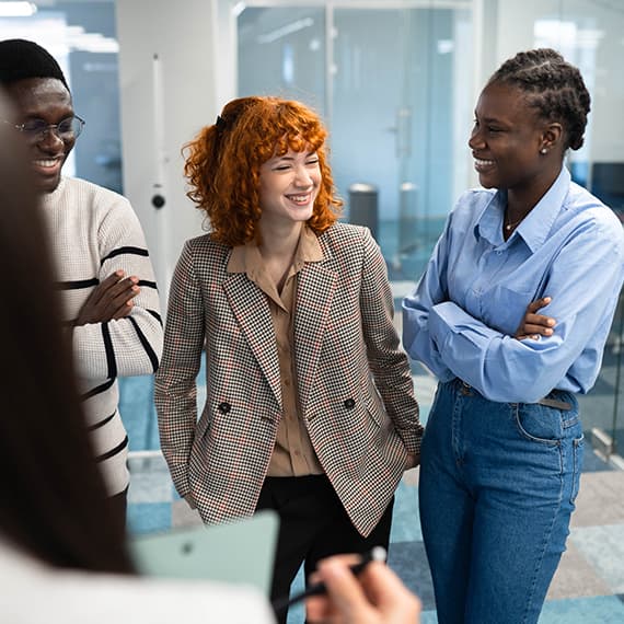 Three diverse professionals smiling and conversing in a modern office with glass walls and bright lighting.
