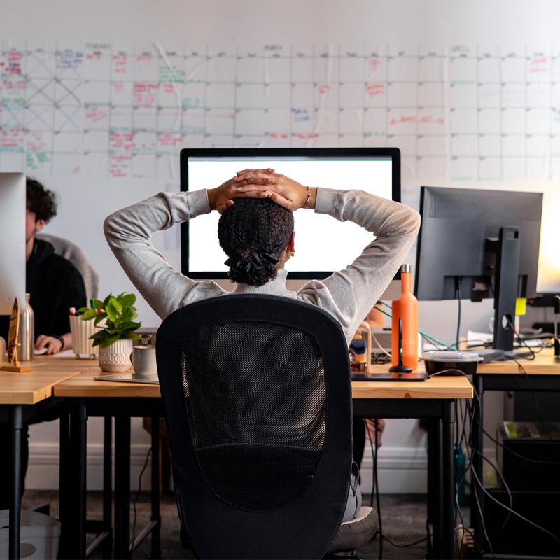 Rear view wide shot of a woman sitting working at a computer in an office. She has her hands behind her head as she stretches, looking at the screen.