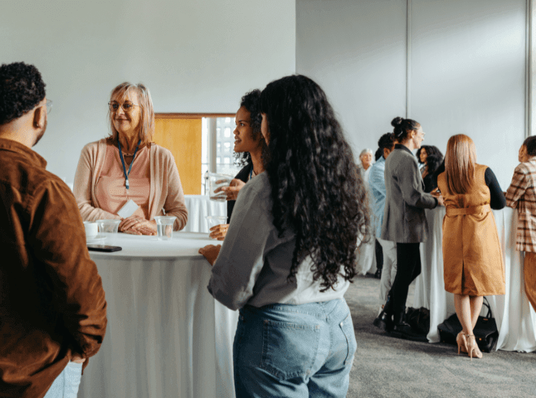 Diverse group of people networking at a professional event, gathered around white cocktail tables in a bright room.