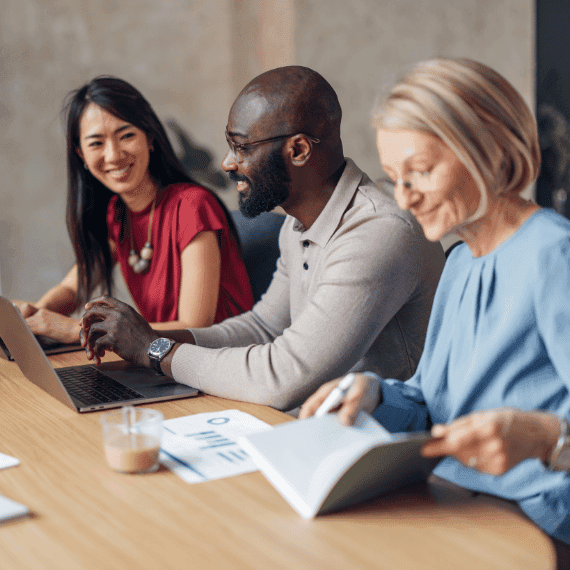 Three diverse professionals collaborating at a wooden table with laptop and documents, smiling during a business meeting.