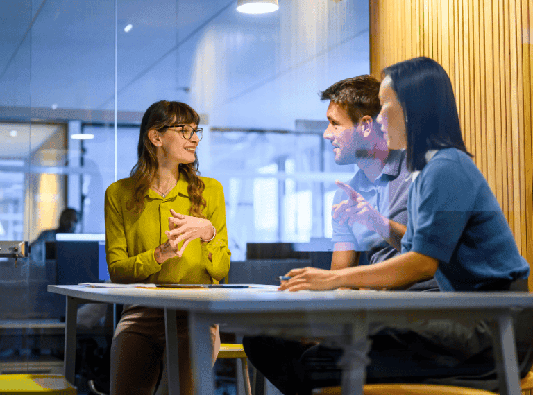 Three professionals having a discussion at a table in a modern office with glass walls and wood paneling.