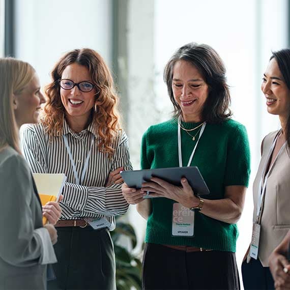 Four professional women smiling and looking at a tablet together in a bright office setting during a business meeting.