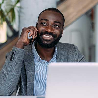Smiling professional in gray blazer and striped shirt talking on phone while working on laptop in bright office space.