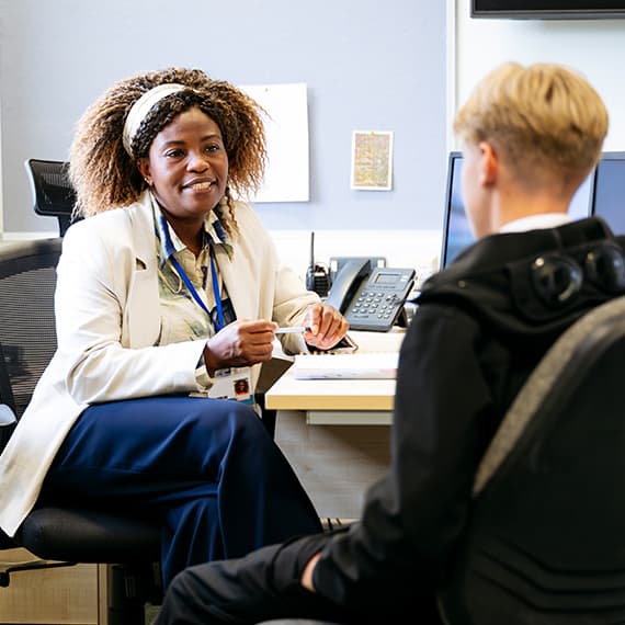 Woman with blue lanyard smiling while meeting with person in office setting with desk and whiteboard visible.