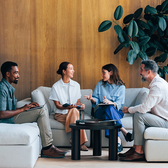 Four professionals sitting on white sofas having a discussion with notebooks against a wooden wall with green plant decor.