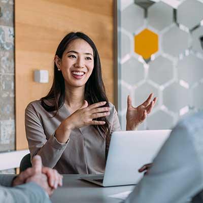 Woman in business meeting gesturing while speaking, with laptop and hexagonal wall design in background.