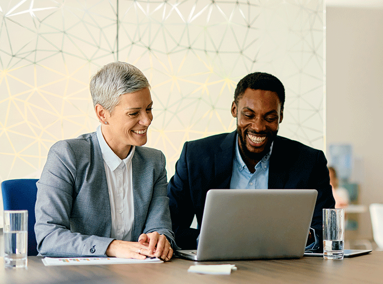 Two smiling business professionals in suits looking at a laptop together at a conference table with geometric wall pattern.