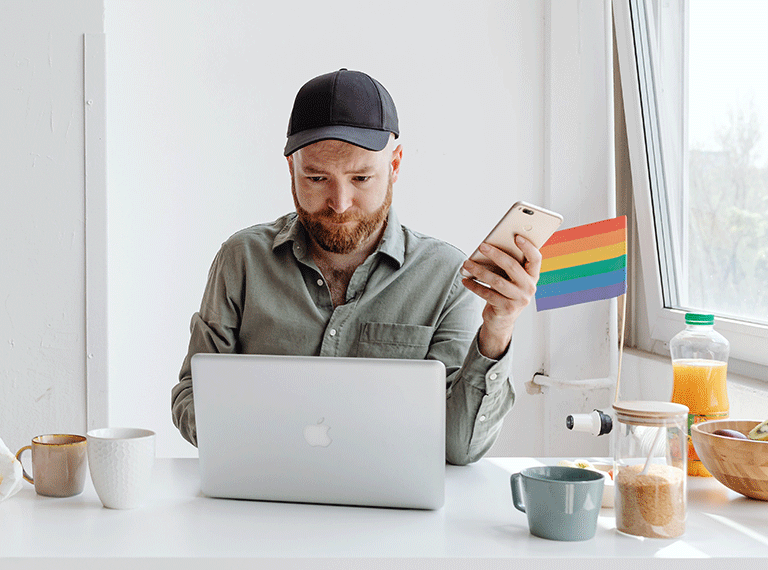 Man wearing black cap and green shirt looking at phone while working on laptop at white desk with coffee and snacks.