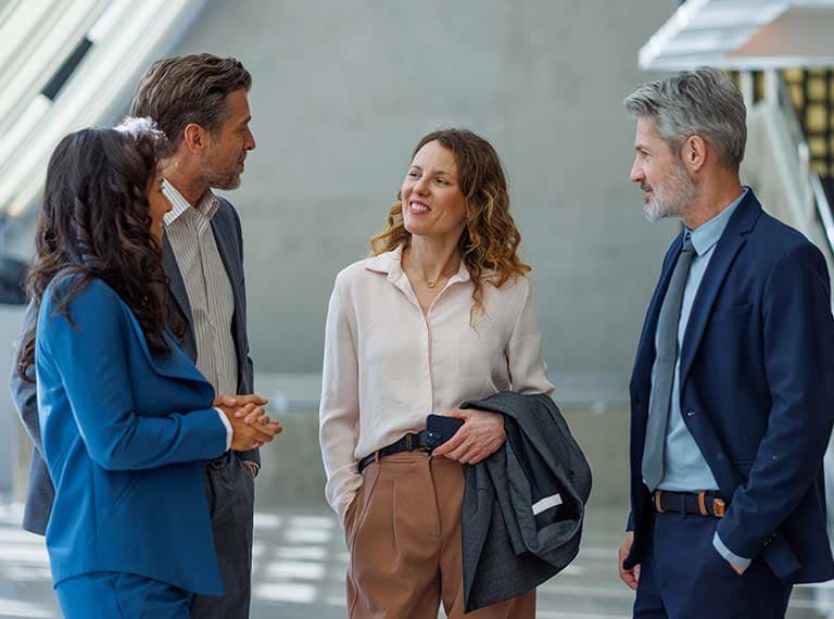 Four business professionals in suits having a friendly conversation in a modern office setting with natural light.
