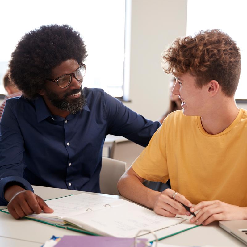 High School Tutor Giving Male Student One To One Tuition At Desk In Classroom.