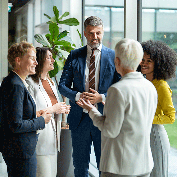 Diverse group of business professionals having a friendly conversation in a bright modern office with large windows and plants