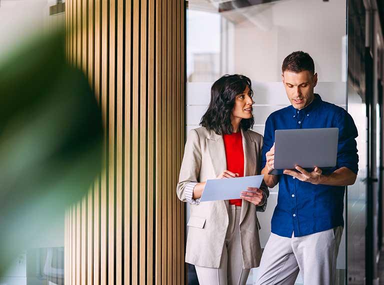 Two professionals in a modern office with wooden slat wall, reviewing documents and laptop together.
