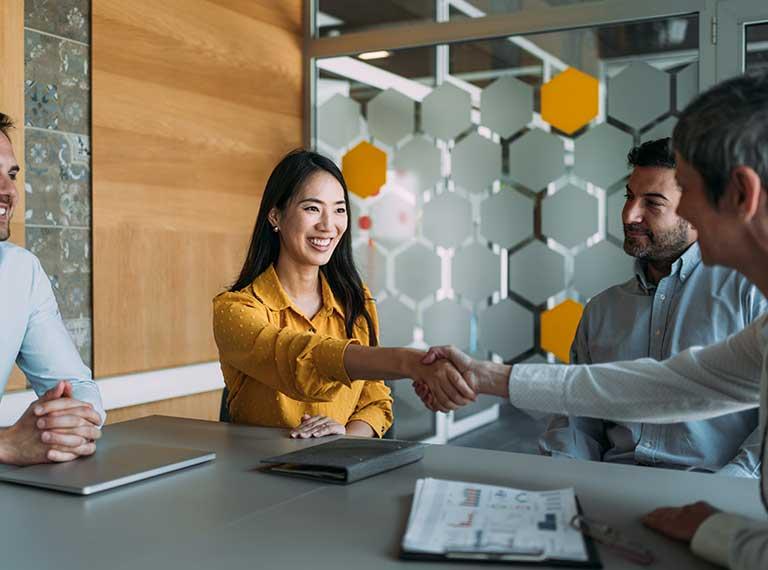 Business meeting in modern office with woman in yellow blouse shaking hands with colleague, hexagonal wall divider visible.