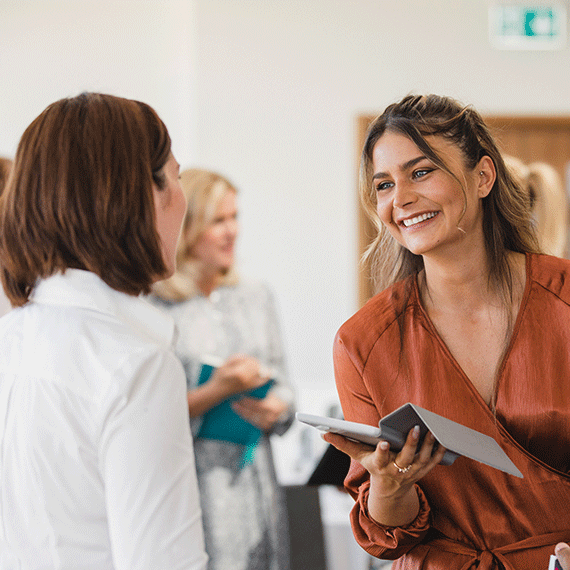 Woman in rust-colored blouse smiling while holding tablet, speaking with person in white shirt in bright office setting.