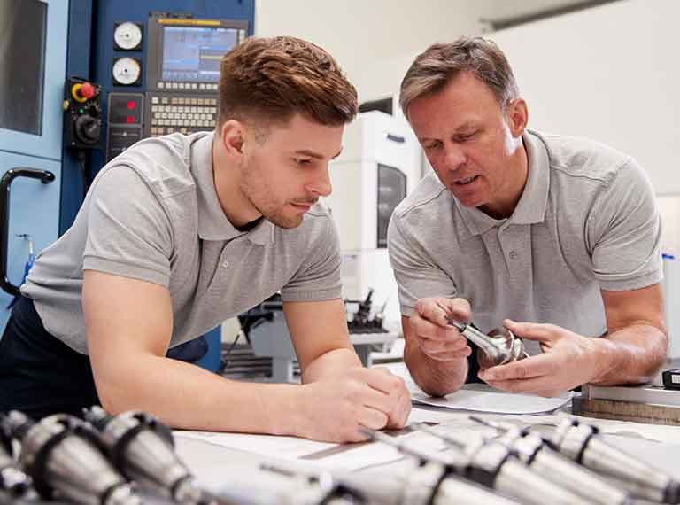Two men in gray polo shirts examining a metal part and documents in a manufacturing facility with CNC machinery visible.