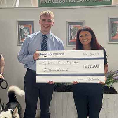 Two people holding a large Reed Foundation cheque for ten thousand pounds, smiling in front of framed posters on a wall.
