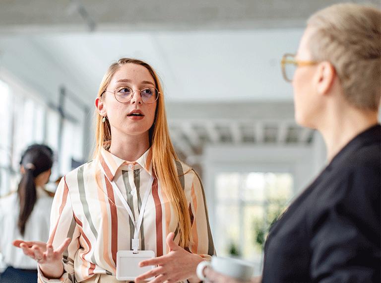 Young woman with glasses in striped shirt speaking to colleague in modern office setting with bright natural light.