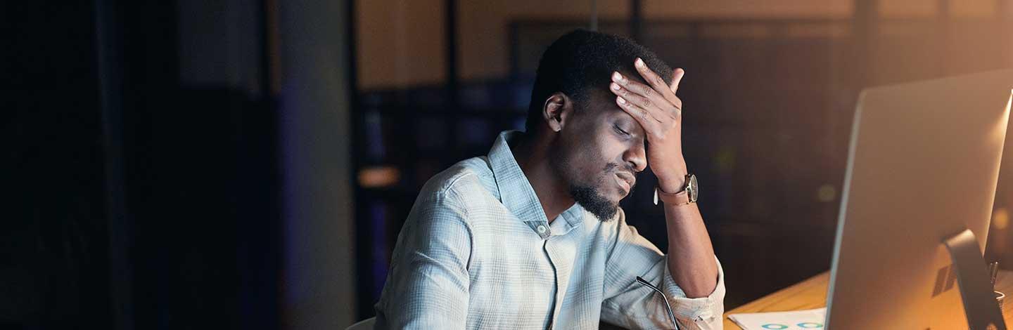 Stressed man in light blue shirt with hand on forehead working late at computer in dimly lit office.