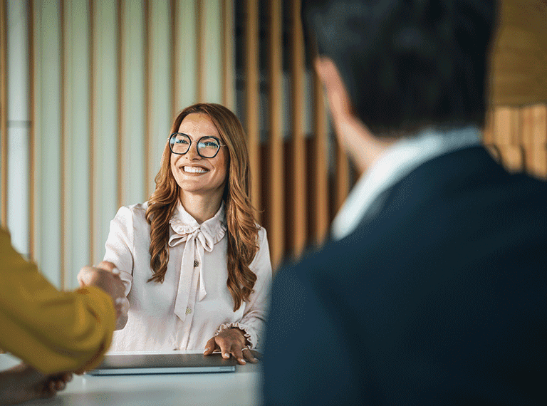 Smiling woman with glasses and long brown hair shaking hands with two people across a desk in an office setting.