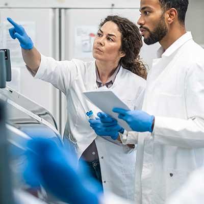 Two scientists in lab coats and blue gloves examining data on a screen in a laboratory setting.