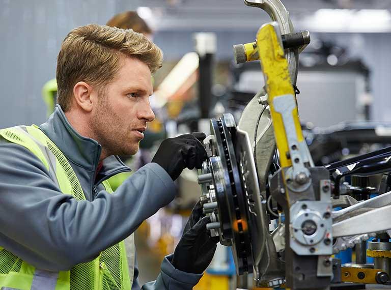 Automotive technician in safety vest examining a brake assembly on a vehicle in a manufacturing facility.
