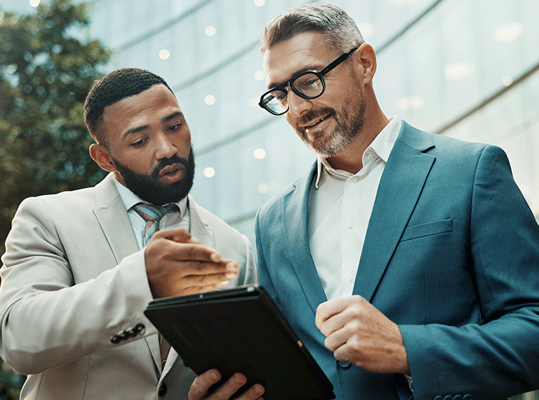 Two businessmen in suits reviewing information on a tablet outdoors in front of a modern glass building.