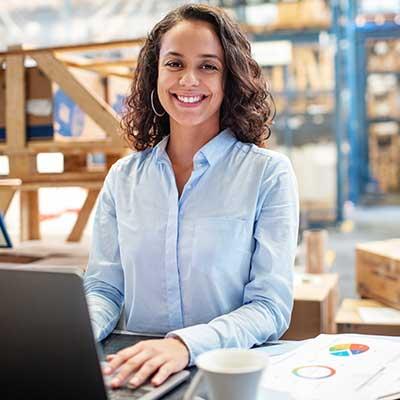 Smiling person in light blue shirt working on laptop in warehouse setting with coffee cup and charts nearby.